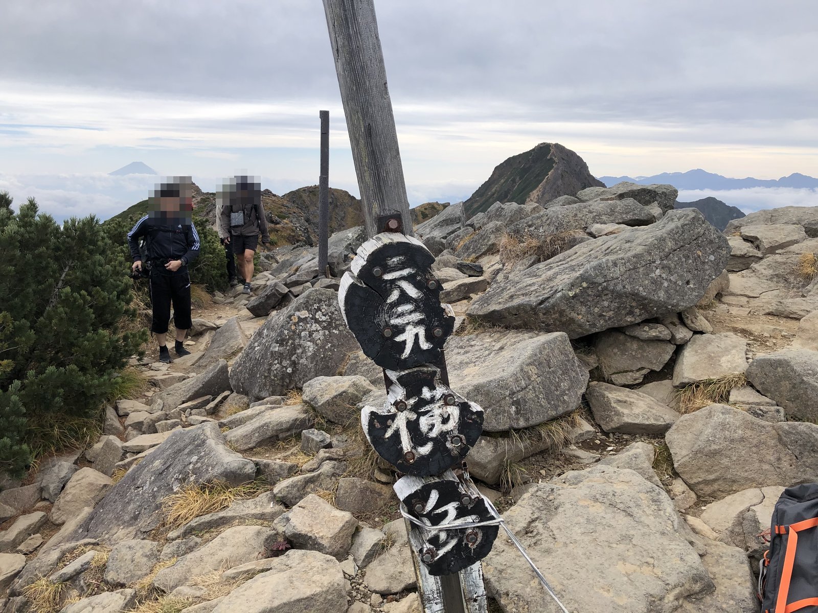 横岳奥ノ院の山頂標識 岩が積み重なる山頂と稜線を行く登山者 南八ヶ岳 ヤマカルテ