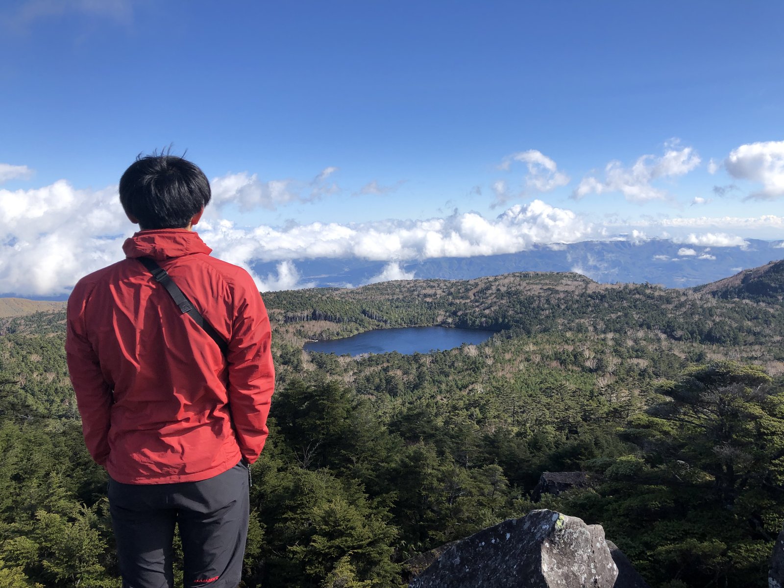 高見石の上から見下ろした森のなかの白駒池と、景色を眺める赤いジャケットの筆者の後ろ姿 北八ヶ岳 ヤマカルテ